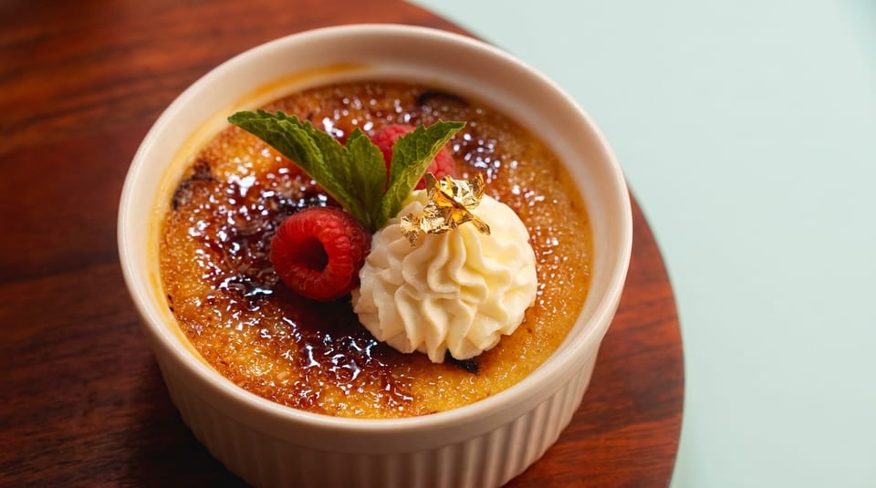 Crème brûlée in a ramekin, topped with whipped cream, gold leaf, raspberries, and mint.