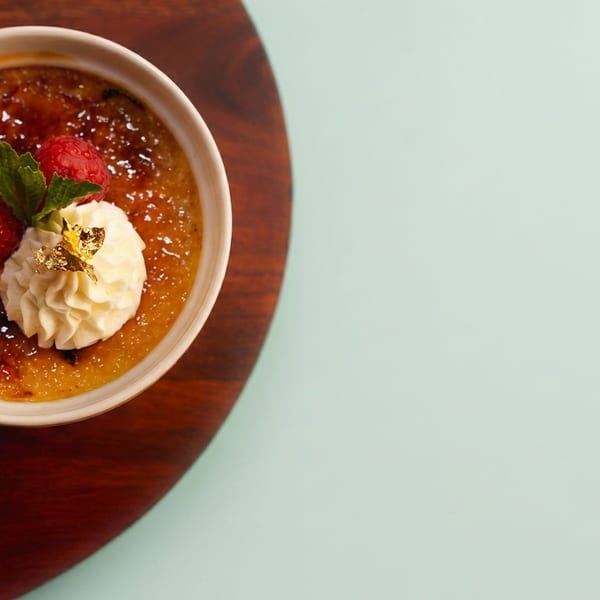 Crème brûlée with raspberries, whipped cream, mint, and gold leaf on a wooden board.