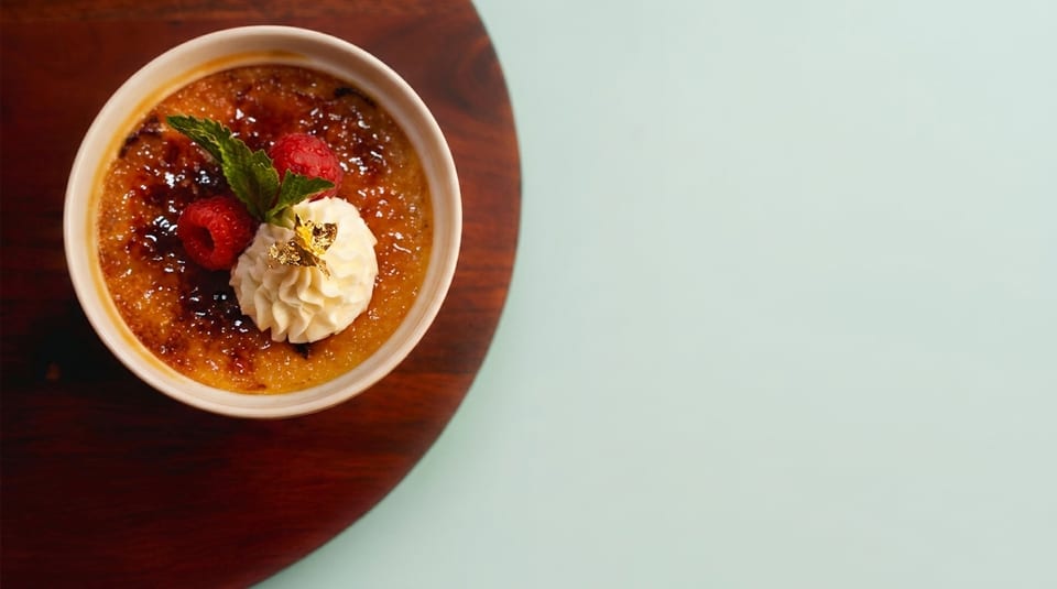Crème brûlée with raspberries, whipped cream, mint, and gold leaf on a wooden board.